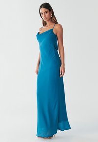 Maksi obleka iz turkiznega satenastega materiala z ovratnikom v obliki ovratne krpe, tankimi nastavljivimi naramnicami in tekočo silhueto. Mehka tekstura in rahlo stransko razporko poudarjata gibanje.