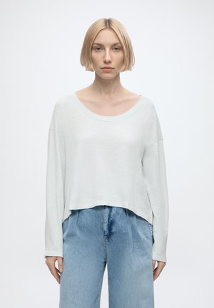 Young woman with short blonde hair wearing a loose white long-sleeve top and high-waisted light blue jeans, standing against a plain white background.