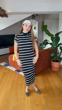 Młoda kobieta uśmiechająca się, ubrana w czarno-białą sukienkę w paski i białą opaskę na głowie, stojąca w salonie przy dużej roślinie doniczkowej.