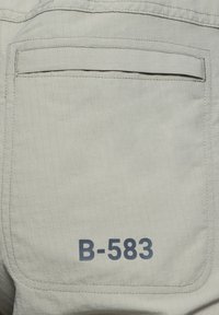 Ljusgrått tyg med rutmönster, med en bakficka och texten "B-583" i mörkblått. Slät textur, ingen synlig hårdvara.