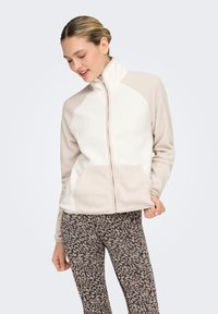Veste en polaire légère couleur crème et taupe avec col montant, fermeture éclair complète et poches avant ; associée à des leggings à motifs.