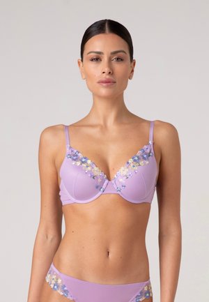 Donna che indossa reggiseno lavanda e mutandine abbinate con ricamo floreale pastello, in piedi davanti a uno sfondo chiaro uniforme.