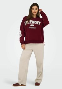 Maroon huppari, jossa on valkoista tekstiä "DETROIT" ja "MICHIGAN." Ominaisuuksiin kuuluu ylisuurikokoinen malli, ribbikäsivarren resorit ja sivuaukot. Beige leveälahkeiset housut.