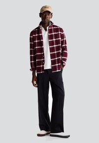 Camicia di flanella a quadri bordeaux e bianca, polo bianca, pantaloni a righe navy, cappellino beige e sneakers bianche con accenti neri e marroni.