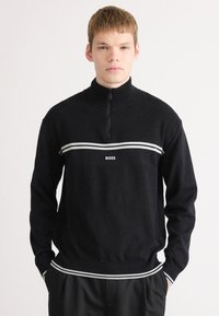 Μαύρο ζακέτα με φερμουάρ και ριμπ υφή, που διαθέτει μια λευκή οριζόντια ρίγα και λογότυπο. Μακριά μανίκια, εφαρμοστό τελείωμα και σχέδιο υψηλού γιακά.