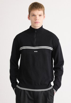 Svart dragkedjesweater med ribbad textur, som har ett vitt horisontellt band och logotyp. Långa ärmar, åtsittande nederkant och design med hög krage.