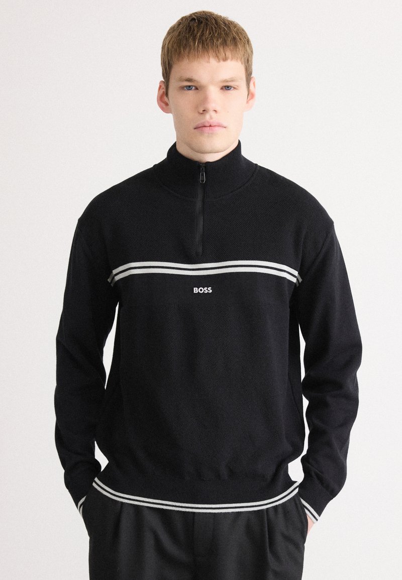 Svetor preto com fecho, com textura canelada, apresentando uma faixa horizontal branca e um logótipo. Manga longa, bainha ajustada e design de colarinho alto.