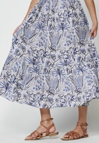 Rochie albă fluidă cu imprimeu botanic albastru, având un tiv în straturi. Sandale maro cu curele și cu ținte aurii completează ținuta.