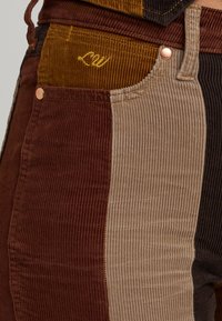 Corduroy pants featuring alternating panels in brown, tan, and dark brown hues. Includes gold embroidery and copper hardware accents.