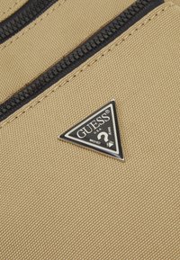 Tissu beige avec une finition texturée, comportant des zips noirs. Un logo triangulaire noir et blanc est affiché de manière proéminente.