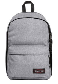 Mochila de tela gris con forma redondeada, cierre de cremallera y bolsillo frontal. Cuenta con un asa superior y un parche de logo en negro y rojo.