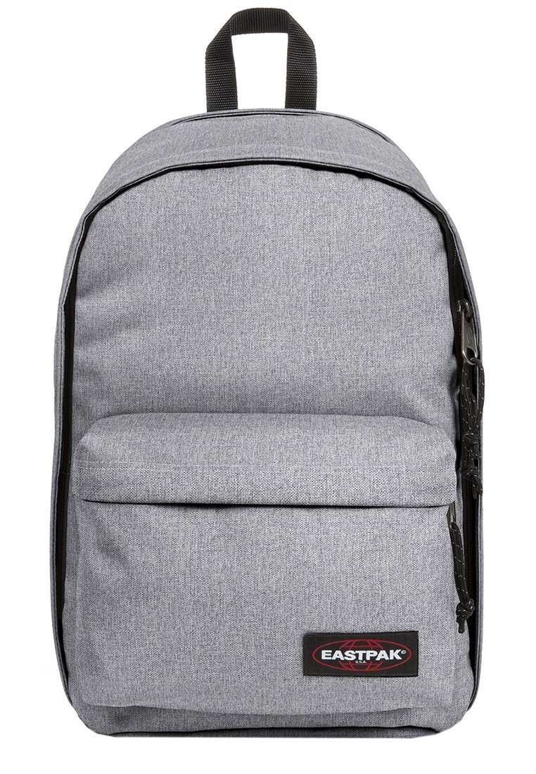 Mochila de tela gris con forma redondeada, cierre de cremallera y bolsillo frontal. Cuenta con un asa superior y un parche de logo en negro y rojo.