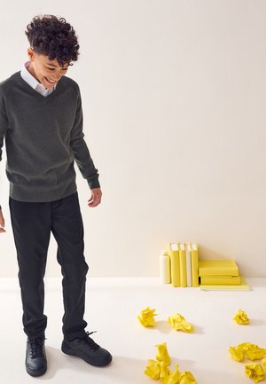 Ung person i en grå sweater og sorte bukser kigger ned og smiler til krøllet gult papir på gulvet nær stablede gule bøger og en flaske.