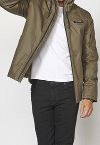 Mężczyzna w oliwkowej kurtce na białej koszuli i czarnych spodniach, poprawiający kurtkę jedną ręką uniesioną w górę.