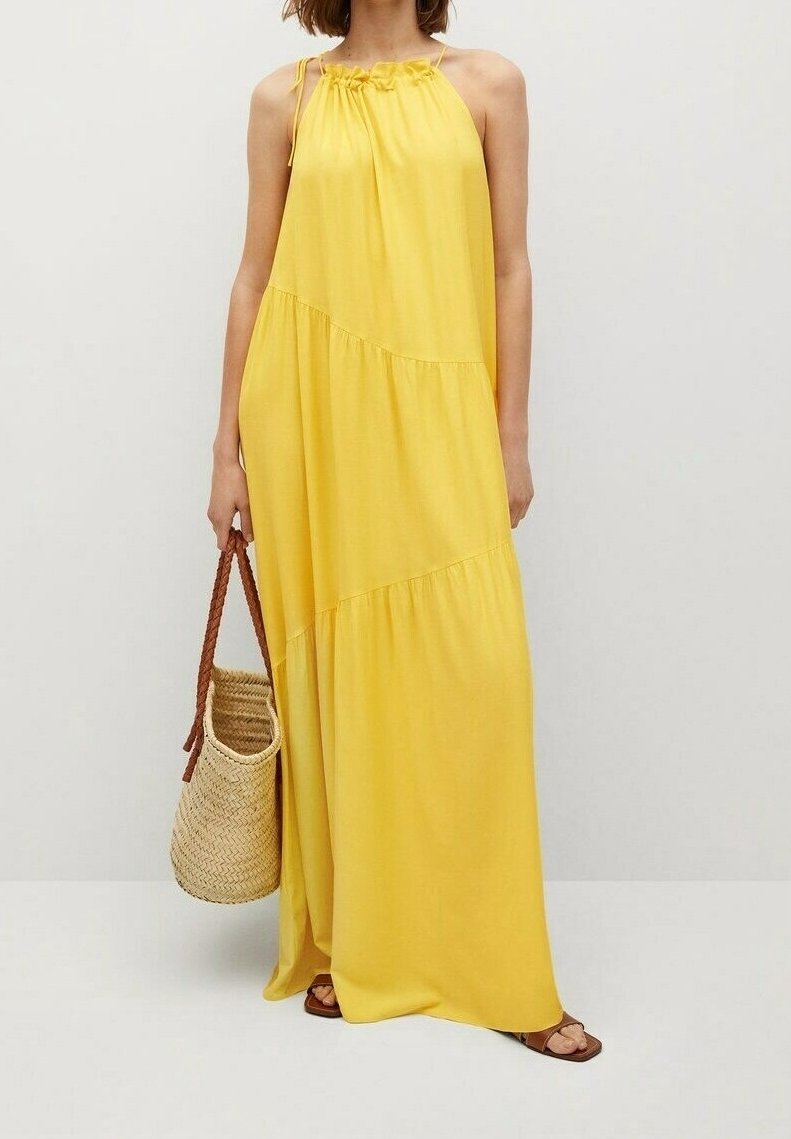 Femme portant une robe maxi jaune sans manches tenant un sac en paille tressée avec des anses marron, debout devant un fond clair uni.