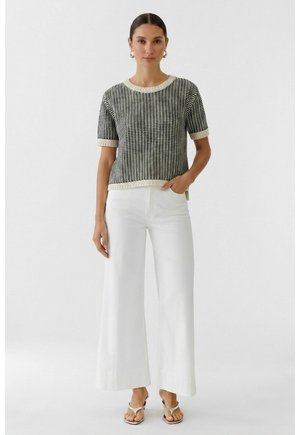 Femme portant un pull à manches courtes à motifs noir et blanc, un pantalon blanc à jambes larges et des sandales blanches à bouts ouverts, debout avec une main dans la poche.
