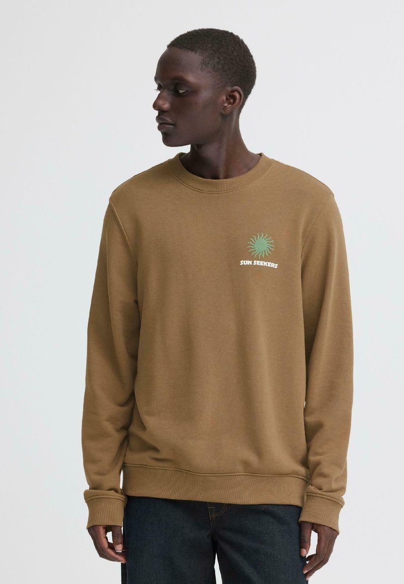 Personne portant un sweat-shirt marron avec un graphique de soleil vert et le texte "Sun Seekers", regardant vers la gauche sur un fond uni.