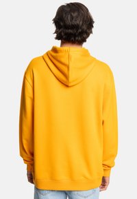 Žltá mikina s kapucňou na šnúrku, rebrovanými manžetami a spodným okrajom, vyrobená z mäkkej tkaniny s hladkým povrchom. Štandardný strih, viditeľné švy.