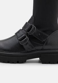 Černá kotníková bota vyrobená z texturovaného materiálu, se dvěma přezkami, silnou podrážkou a kulatou špičkou, s designem na podrážce.