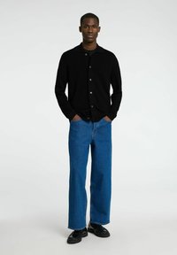 Svart strikket cardigan med spiss krage og frontknapper, sammen med blå vide bukser og svarte sko, står mot en ensfarget bakgrunn.