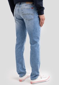 Mees kannab helesiniseid sirge säärega teksapükse ja valgeid tossusid, seisab parema käega reiel toetudes, tavalise taustaga.