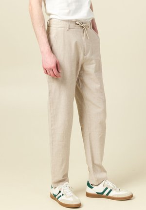 Mann trägt beige Kordelzughose, weißes T-Shirt und weiße Sneakers mit grünen Streifen, steht vor einem einfarbigen Hintergrund.