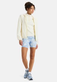 Light yellow zip-up jacket with elastic cuffs, paired with a white ribbed top and blue patterned shorts. Completed with blue sneakers.