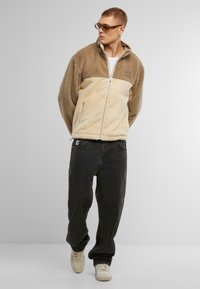Jachetă din fleece în nuanțe de bej și crem, cu fermoar pe față, guler înalt și material texturat. Asortată cu pantaloni închisi la culoare și adidași deschiși.