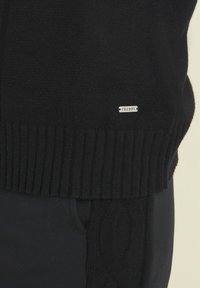 Pull noir en matériau doux et texturé avec un ourlet côtelé. Il présente une petite étiquette en argent avec le nom de la marque "Freddy" sur le côté.