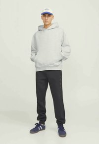 Jovem de pé com as mãos nos bolsos, vestindo um hoodie cinza claro, calças de treino pretas, ténis azuis e um boné branco com azul.