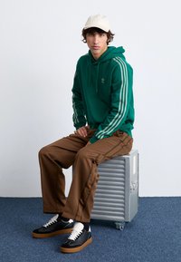 Green hoodie with two vertical stripes, brown track pants with side stripes, and black sneakers with white accents, all displayed on a metal crate.