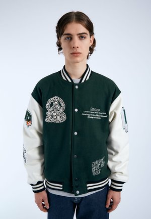 Veste universitaire verte et crème avec des designs brodés, un col rayé, des boutons-pression et des accents en texte. Tissu texturé avec des coutures visibles.