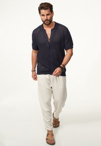 Marineblaues kurzärmeliges Strickhemd mit offenem Gewebe, Knopfverschluss und Kragen, kombiniert mit locker sitzenden, cremefarbenen Hosen und Sandalen.