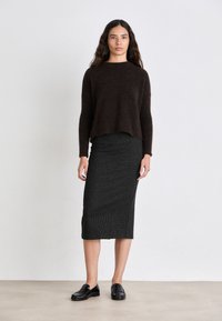 Brauner Strickpullover, taillierter Schnitt. Schwarze Falte mit vertikalen weißen Streifen. Model trägt schwarze Slipper und steht vor einem neutralen Hintergrund.