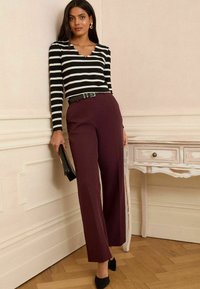Haut à rayures noir et blanc à manches longues avec un col festonné, associé à un pantalon large bordeaux et des talons noirs, tenant une pochette noire.