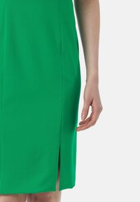 Grünes Kleid mit glatter Textur, das einen seitlichen Schlitz und eine taillierte Silhouette aufweist. Nahaufnahme einer Hand in der Nähe des Saums des Kleides.