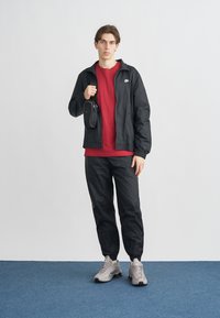Black zip-up jacket, red t-shirt, black joggers, and gray sneakers. Features Nike logo on jacket, textured fabric, and a small bag.