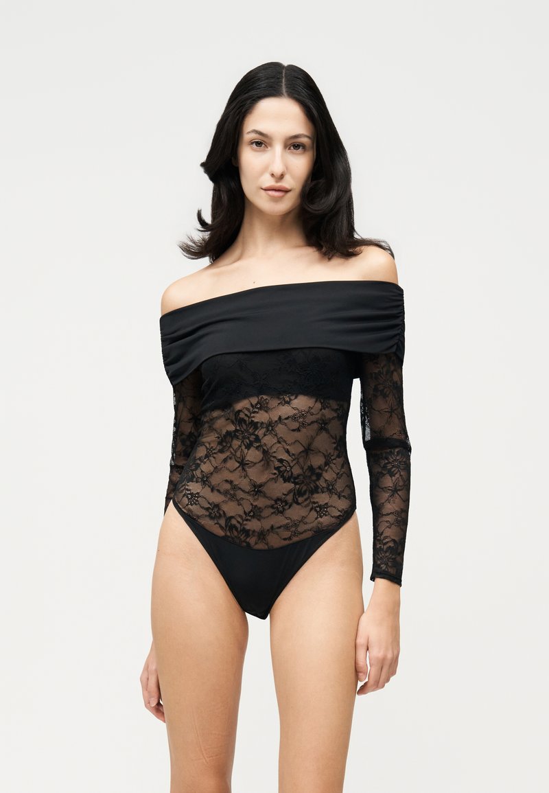 Une femme porte un body noir à épaules dénudées, avec des manches et un buste en dentelle florale transparente, sur un fond clair uni.