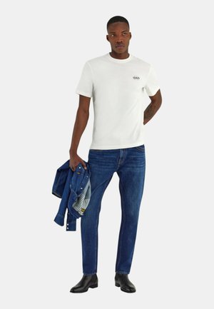 Uomo con t-shirt bianca a maniche corte, jeans blu e stivali neri, che tiene in mano sinistra una giacca di denim blu, in piedi su sfondo bianco.