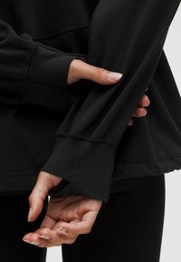 Čierna mikina s dlhými rukávmi, s rebrovanými manžetami, jemnou textúrou a diskrétnymi detailmi šitia. Detailný záber rúk držiacich látku.