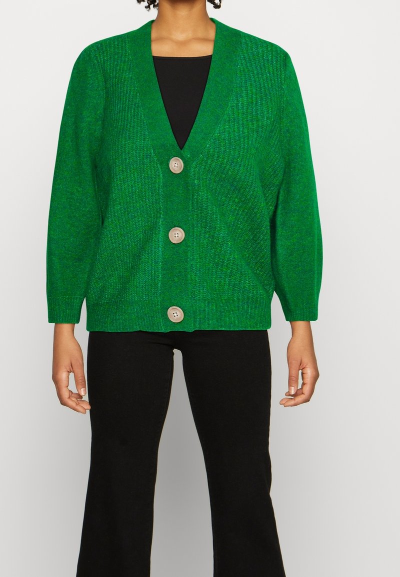 Personne portant un cardigan vert vif avec trois gros boutons beiges, un haut noir et un pantalon noir, debout devant un fond uni.