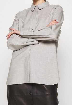 Person iført en lysegrå langærmet skjorte med knapper og krave, armene krydset, og et sort lædernederdel mod en hvid baggrund.