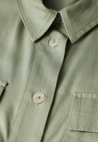 Camicia a maniche corte di colore verde chiaro, realizzata in tessuto morbido. Presenta un colletto a punta, zwei bottoni e una tasca sul petto con cuciture visibili.