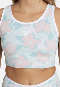 Květinový vzorovaný crop top s světle růžovými a tyrkysovými květinami na bílém podkladu. Má kulatý výstřih a kontrastní bílý lem.