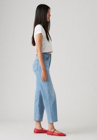 Jauna sieviete stāv sānis, ģērbta baltā īsajā T-kreklā, gaismas zilonī augstās jostās džinsos un sarkanos plakano zābakos.