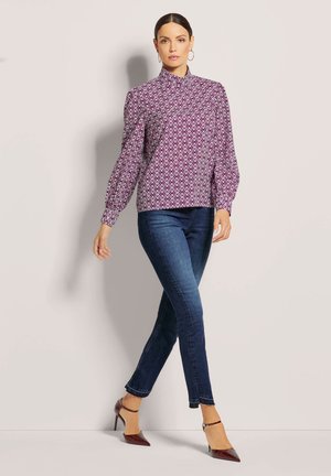 Lila gemusterte Bluse mit hohem Ausschnitt und Ballonärmeln; kombiniert mit dunkelblauen Jeans und burgunderfarbenen spitzen Pumps. Glatte, eng anliegende Texturen.