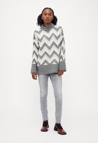 Pullover grigio e bianco con motivo a zig zag e collo alto, abbinato a jeans grigi chiari e scarpe con tacco quadrato rosse e lucide.