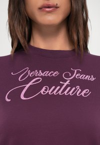 Ljubičasta pamučna majica s istaknutim sjajnim ružičastim logotipom "Versace Jeans Couture" u kurzivnom tisku na prednjoj strani.