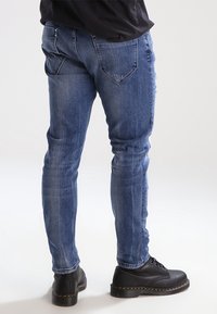 Slim-fit modré džíny s klasickými zadními kapsami a jemným vyblednutím, doplněné černými šněrovacími botami s žlutým prošíváním.
