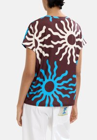 Brązowy t-shirt z krótkim rękawem, z dużymi niebieskimi i kremowymi wzorami w kształcie promieni słonecznych. Ma luźny krój i małą metkę u dołu.
